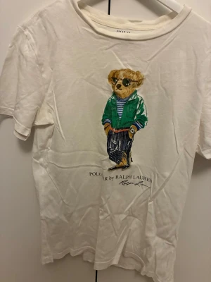 Vit t-shirt med Polo Bear från Ralph Lauren - Vit t-shirt från Ralph Lauren med ikoniskt Polo Bear-motiv på framsidan. Björnen har en grön jacka, randig tröja och solglasögon. T-shirten har rund hals och korta ärmar, perfekt för en avslappnad och cool look.