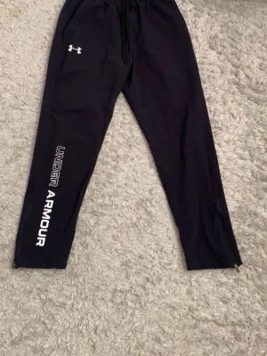  Under Armour byxor - Snygga svarta träningsbyxor från Under Armour med vit logga på låret och stort tryck längs benet. Byxorna har dragsko i midjan, fickor och dragkedjor vid bensluten. Perfekta för gymmet eller löprundan.