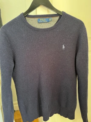 Mörkblå stickad tröja Polo Ralph Lauren - Mörkblå stickad tröja från Polo Ralph Lauren med rund halsringning och diskret broderad logga på bröstet. Tröjan har långa ärmar och ribbade muddar vid ärmslut och nederkant. Klassisk och stilren modell i mjukt material.