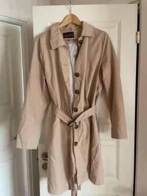 Beige trenchcoat från Wera Stockholm - Stilren beige trenchcoat från Wera Stockholm med knappar framtill och knytskärp i midjan. Klassisk krage och långa ärmar. Tunnare material, perfekt för vår och höst. Fodrad med randigt innerfoder för en snygg detalj. Storleken är 44 men den är lite mindre i storleken