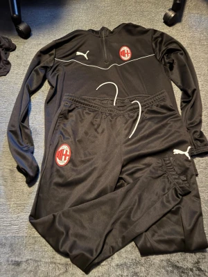 AC Milan svart träningsset Puma - Svart träningsset med långärmad tröja och byxor från Puma, prydd med AC Milan-logga på både tröja och byxa. Setet har vita detaljer och dragkedja vid halsen. Perfekt för fotbollsträning eller chill. Materialet är mjukt och andas.