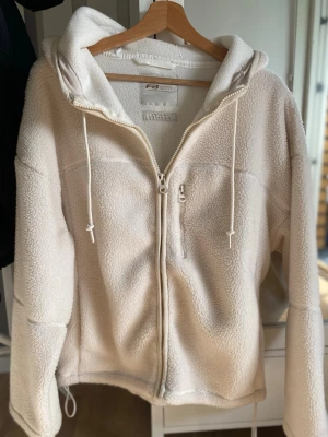 Vit teddyhoodie från FB Sister - Mysig vit teddyhoodie från FB Sister med dragkedja framtill och stor huva. Tillverkad i fluffigt, mjukt teddy-material som ger en avslappnad och trendig look. Perfekt för dig som gillar oversized och bekväma plagg. Justerbar nederkant med dragsko.