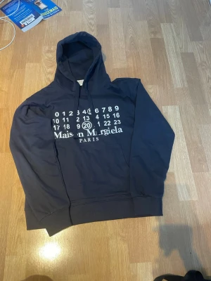 Mörkblå Maison Margiela hoodie - Cool mörkblå hoodie från Maison Margiela Paris med ikoniskt tryck av siffror på bröstet. Hoodien har huva med dragsko och ribbade muddar. Materialet är mjukt och bekvämt, perfekt för en avslappnad och trendig look.
