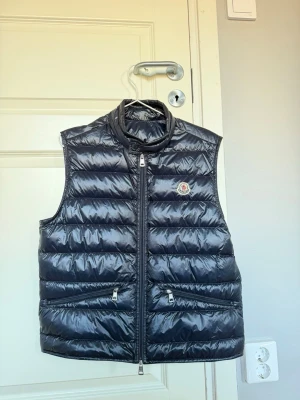 Mörkblå  dunväst från Moncler - Snygg svart dunväst från Moncler med glansig finish och quiltad design. Västen har två dragkedjeförsedda fickor framtill och en hög krage. Moncler-loggan sitter på bröstet. Perfekt för lager-på-lager och streetwear-stil.