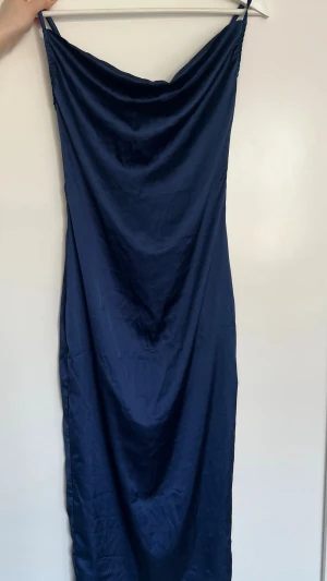 Marinblå långklänning i satin - Snygg marinblå långklänning i glansigt satinmaterial med smala axelband och draperad urringning. Klänningen har en elegant och enkel design som ger en lyxig känsla. Perfekt för dig som vill sticka ut med stilren look.