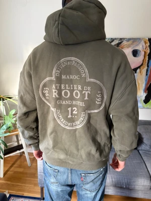 Olivgrön hoodie - Säljer en olivgrön hoodie från Atelier de Root med stor tryckt logga på ryggen och mindre tryck framtill. Hoodien har huva och en stor magficka. 