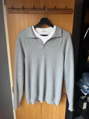 Ljusgrå tröja från John Henric  - Säljer en stilren ljusgrå stickad tröja med krage och v-ringning. Tröjan har lång ärm och är ribbstickad för en snygg struktur. Perfekt att bära över en t-shirt för en avslappnad men ändå uppklädd look.