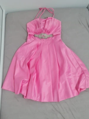 Det kan vara s Rosa kort klänning med strassdetalj - Supersöt rosa kort klänning i glansigt material med tunna axelband och snygg strassdetalj i midjan. Klänningen har v-ringad överdel och ett svepande kjolparti som ger en elegant look. Perfekt för dig som vill sticka ut!