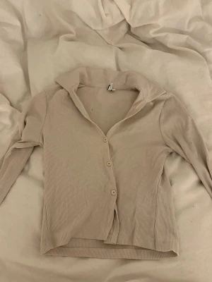 Beige ribbad cardigan med krage - Trendig beige ribbad cardigan med tre knappar framtill och bred krage. Långärmad modell i mjukt, stretchigt material som passar perfekt till lager på lager. Enkel och stilren design som är lätt att matcha.