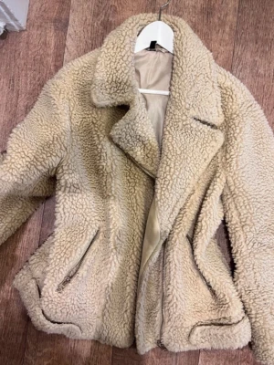Beige teddyjacka med dragkedjor - Säljer en fluffig beige teddyjacka med bred krage och två snedställda dragkedjefickor framtill. Jackan har en klassisk bikerinspirerad look och är fodrad på insidan för extra värme. Perfekt för dig som vill ha en mysig och trendig jacka i höst och vinter.
