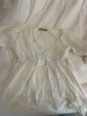 Vit omlottblus med volangdetaljer - Lång i längden. Säljer en luftig vit blus med omlott-skärning och fina volangdetaljer längs midjan och nederkanten. Blusen har v-ringning och trekvartsärm, vilket ger en avslappnad och romantisk look. Perfekt att styla med jeans eller kjol. 