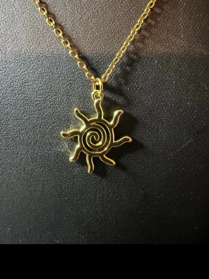 Guldigt halsband med solhänge - Unikt halsband i guldfärgad metall med ett solformat hänge. Hänget har en spiral i mitten och solstrålar som sticker ut, vilket ger en cool och bohemisk vibe. Perfekt för dig som gillar statement-smycken och vill sticka ut lite extra.