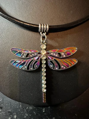 Halsband med trollslända och pärlor - Unikt halsband med svart läderrem och en stor trollslända som hänge. Trollsländan har färgglada vingar i lila, blått, orange och silver samt gnistrande pärlor längs kroppen. Perfekt statement piece för dig som gillar färg och detaljer.