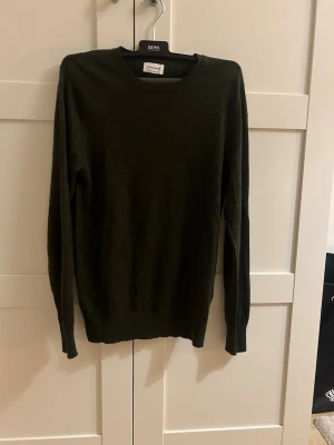 Mörkgrön stickad tröja från A days march - En stilren mörkgrön stickad tröja från A days march med rund halsringning och långa ärmar. Tröjan har en smal passform och ribbade muddar vid ärmslut och nederkant. Perfekt för dig som gillar klassisk och enkel stil.