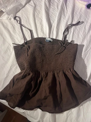 Brun babydoll topp från American Eagle - Säljer en brun smockad topp från American Eagle med justerbara axelband som knyts. Toppen har en stretchig överdel och en luftig, utställd nederdel. Perfekt för sommaren och enkel att styla med jeans eller kjol. Passar S! Helt ny, aldrig använt 