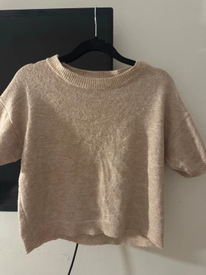 Beige kortärmad stickad tröja - Säljer en beige, kortärmad stickad tröja med rund hals och ribbade kanter. Tröjan har en avslappnad passform och är perfekt för lager på lager. Materialet känns mjukt och bekvämt, troligen en blandning av bomull och syntet. Från SHEIN i storlek S