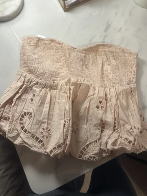 Beige axelbandslös tubtopp med broderi - Söt beige tubtopp med smock upptill och volangig nederdel. Toppen har fina broderade detaljer och hålmönster som ger en bohemisk känsla. Perfekt för varma dagar och enkel att styla till jeans eller kjol. Från Zara - S