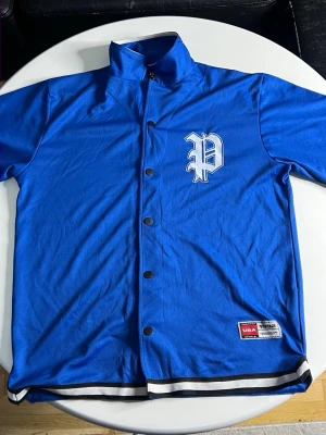 Blå basebolltröja med tryck - Snygg blå basebolltröja med tryckknappar och ett vitt P-tryck på bröstet. Tröjan har svartvita ränder längst ner och står ut med sin sportiga look. Tillverkad i ett lätt och ventilerande material, perfekt för träning eller streetwear.