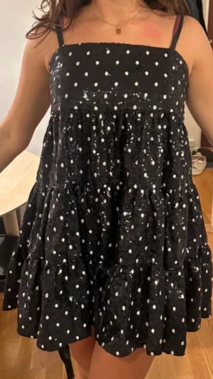 Svart kort prickig klänning paletter - Supersöt svart kort klänning med vita prickar och tunna axelband. Klänningen har en volangig och luftig passform som ger en lekfull vibe. Perfekt för varma dagar och när du vill sticka ut lite extra. Paletter sockersöt