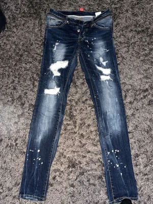 Mörkblå distressed jeans Dsquared2 - Snygga mörkblå jeans från Dsquared2 med slitningar, hål och vita färgstänk för en trendig, edgy look. Smal passform och klassisk femficksdesign. Tillverkade i Italien av denim med autentisk känsla.