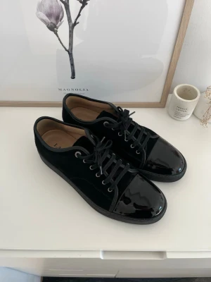 Lanvin cap toe  - Stilrena svarta sneakers från Lanvin med snygg kombination av blankt lack och mocka. Klassisk låg modell med svarta skosnören och diskret logga på insidan. Perfekta för dig som gillar en clean och lyxig look. Använda endast 1 gång!