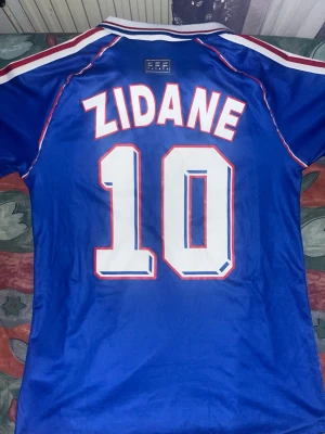 Frankrike Zidane tröja Adidas VM 98 - Blå fotbollströja från Adidas med ZIDANE och nummer 10 på ryggen, inspirerad av Frankrikes VM 1998. Tröjan har vita och röda detaljer, vit krage och broderad Adidas-logga framtill. Perfekt för fotbollsfans och samlare.