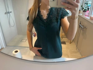 Mörkgrön blus med spetsdetaljer - Supersnygg mörkgrön blus med v-ringning och spetsdetaljer vid hals och ärmar. Blusen är ärmlös och har en feminin, elegant look. Perfekt att styla med jeans eller kjol för en trendig outfit.