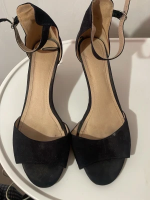 Svarta mocka-klackar med ankelrem - Säljer ett par stilrena svarta klackar med öppen tå och ankelrem. Skorna har en klassisk design med klack och är tillverkade i mocka för en mjuk och lyxig känsla. Perfekta för dig som vill ha en enkel men elegant look.
