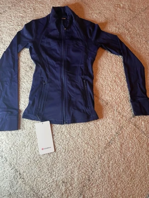 Marinblå träningsjacka Lululemon - Slimmad marinblå träningsjacka från Lululemon med hel dragkedja framtill och två dragkedjeförsedda fickor. Tillverkad i ett stretchigt funktionsmaterial som andas, perfekt för träning eller löpning. Hög krage och långa ärmar för extra komfort.
