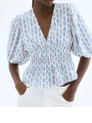 Vit och blå blus med puffärm - Superfin blus i vitt med blått blommigt mönster, v-ringad och med smockad midja. Puffiga korta ärmar ger en trendig look och toppen är lätt croppad. Perfekt för dig som gillar en söt och somrig stil. Säljer min Topp från hm som jag aldrig använt 🤍🩵