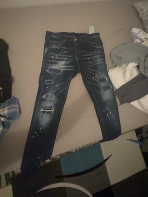 Mörkblå distressed jeans från Dsqaured - Säljer ett par mörkblå jeans från Dsqaured slitningar och trasiga detaljer på benen. Jeansen har en klassisk femficksmodell och coolt spräckligt mönster. Perfekt för dig som gillar en edgy och trendig look.