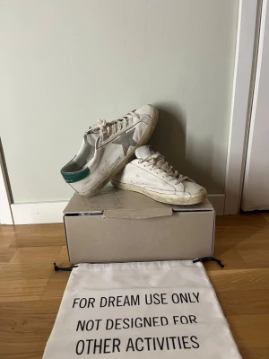 Golden Goose vita sneakers med grön häl - Golden Goose sneakers i vitt läder med grå stjärndetalj på sidan och grön häl. Klassisk låg modell med slitna detaljer, vita skosnören och beige sula. Perfekta för en avslappnad och trendig streetstyle-look.