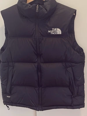 Svart dunväst från The North Face - Svart dunväst från The North Face med hög krage, dragkedja och två fickor. Västen har en puffig design och logga på bröstet