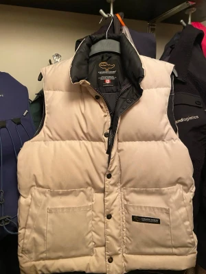 Beige dunväst från Canada Goose - Snygg beige dunväst från Canada Goose. Höger ficka är här en lite skada men kan sys ihop enkelt. 