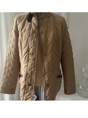 Beige quiltad jacka med dragkedja Abacus  - Snygg beige quiltad jacka med dragkedja och tryckknappar framtill. Jackan har två fickor med silverfärgade dragkedjor och en hög krage. Perfekt för dig som gillar stilren och klassisk look.