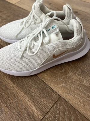 Vita Nike sneakers med mesh - Snygga vita sneakers från Nike med ovandel i ventilerande mesh och klassisk swoosh i beige på sidan. Skorna har vita snören och en bekväm, lätt sula som passar perfekt till streetwear-looken. Diskreta detaljer i turkos på plösen och snörningen.