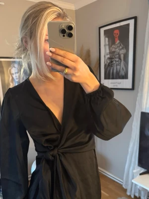 Svart omlottklänning med knyt - Svart kort omlottklänning med knyt i midjan och v-ringad hals. Klänningen har långa, puffiga ärmar och en elegant, enkel design. Perfekt för dig som gillar stilrena plagg med en twist.