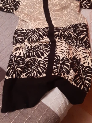 Svartvit mönstrad kortklänning - Kort klänning med svartvitt bladmönster och markerade svarta linjer framtill. Klänningen har rund hals, korta ärmar och ett vitt bälte i midjan. Materialet känns mjukt och stretchigt, troligen bomull eller polyester.