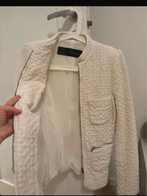 Vit bouclé cardiganjacka från Zara - Stilren vit cardiganjacka från Zara Basic i bouclétyg. Jackan har två framfickor med lock, rund halsringning och dragkedja framtill samt vid fickorna. Perfekt för en chic och trendig look.