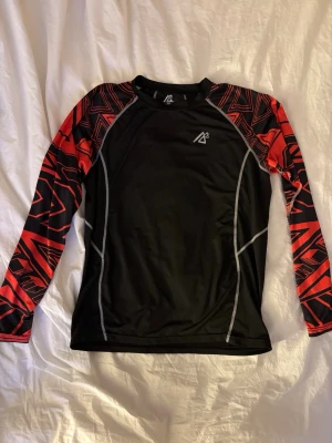 Svart compression shirt med röda mönstrade ärmar - Snygg långärmad sporttröja från Mountain Warehouse i svart med röda och svarta grafiskt mönstrade ärmar. Tröjan har vita kontrastsömmar och är tillverkad i ett stretchigt funktionsmaterial som passar perfekt till träning eller löpning. Storlek XXL men sitter mer som en M/L 