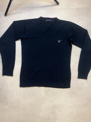 Svart V-ringad tröja från Gant - Obs! det står storlek S men den passar 152-158!!!!!Klassisk svart V-ringad tröja från Gant med broderad logga på bröstet. Tröjan har långa ärmar, ribbade muddar och är tillverkad i ett mjukt stickat material. Perfekt för en stilren och tidlös look. Ganska okej skick skriv om det är nåt du funderar över 
