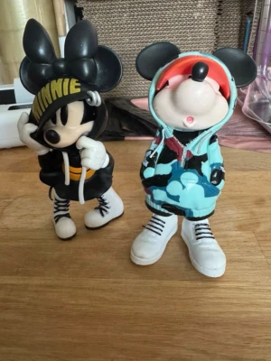 Coola Mickey & Minnie Mouse figurer i streetwear 15cm - Två dekorativa Musse & Mimmi figurer inspirerade av streetwear-stil. Ena figuren har svart hoodie med texten 'MINNIE' i gult och vita sneakers, den andra har blå hoodie med mönster, röd keps och vita sneakers. Perfekt som dekoration eller samlarpryl. Ca 15cm höga
