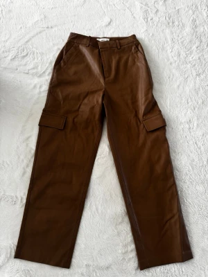 Bruna cargobyxor i fuskskinn - Snygga bruna cargobyxor i fuskskinn med raka ben och stora fickor på sidorna. Byxorna har hällor för skärp och klassisk cargomodell med modern twist. Perfekta för dig som vill sticka ut med en trendig look.