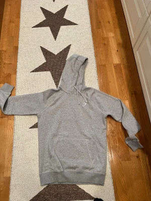 Grå hoodie från Gymshark - Säljer en stilren grå hoodie från Gymshark i storlek M. Hoodien har klassisk känguruficka, dragsko i huvan och ribbade muddar vid ärmslut och nederkant. Tillverkad i mjukt bomullsmaterial som är skönt att ha på sig. Perfekt för dig som gillar enkel och clean stil.
