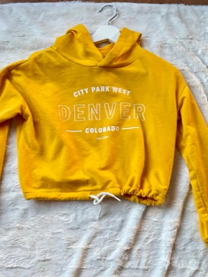 Gul cropped hoodie Denver-print - Gul hoodie med huva och vitt tryck framtill där det står 'City Park West Denver Colorado'. Croppad modell med dragsko i midjan och långa ärmar. Perfekt för en avslappnad och trendig look. Mjuk bomullskänsla och ribbade muddar vid ärmslut.