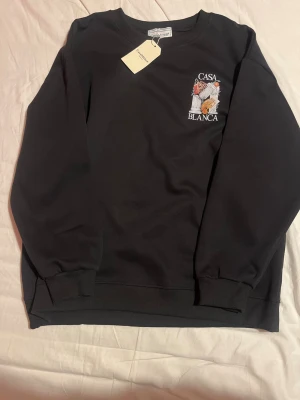 Svart sweatshirt från Casablanca - Snygg svart sweatshirt från Casablanca med broderad logga och färgglatt motiv på bröstet. Tröjan har rund halsringning, ribbade muddar och är gjord i mjukt bomullsmaterial. Perfekt för en chill och stilren look. Den är helt ny med prislapp och aldrig använt den eftersom den är alldeles för stor. Det står att den är XXL men den passar även L-XL då den passar folk från 180-185cm lång