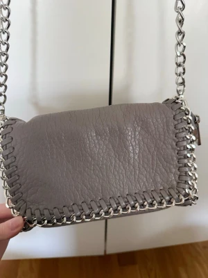 Tiamo Pricilla väska  - liten axelremsväska från Tiamo i modellen pricilla, grå/beige. Knappt använd och inga slitningar repor mm. Kan även bäras jättefint som en clutch om man tar av kedjan. Axellänk 105 cm. Längd 21 cm, höjd 14 cm, bredd 5 cm.