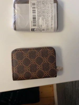 Brun mönstrad plånbok med dragkedja - Snygg brun plånbok med grafiskt mönster och guldfärgad dragkedja. Plånboken har flera fack på insidan för kort och sedlar. Tillverkad i konstläder med stilren design som passar perfekt i fickan eller väskan. Du köper din oöppnade😊