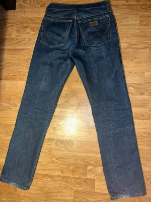 Blå raka jeans från Wrangler - Klassiska mörk denim Wrangler Texas med raka ben och snygga kontrastsömmar. Bakfickorna har det ikoniska W-mönstret och en liten Wrangler-logga. Storlek 30/34.