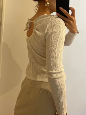 Vit blus med detaljer  - Trendig vit topp med lång ärm och snygg struktur i tyget. Toppen har en öppen rygg med knytband upptill som ger en unik detalj. Även liten puff vid axlarna 